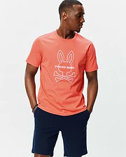 Mens Craig Graphic Tee, a classic fit coral jersey t-shirt featuring a large white outline Psycho Bunny skull and crossbones logo with text prominently on the center chest. (6)