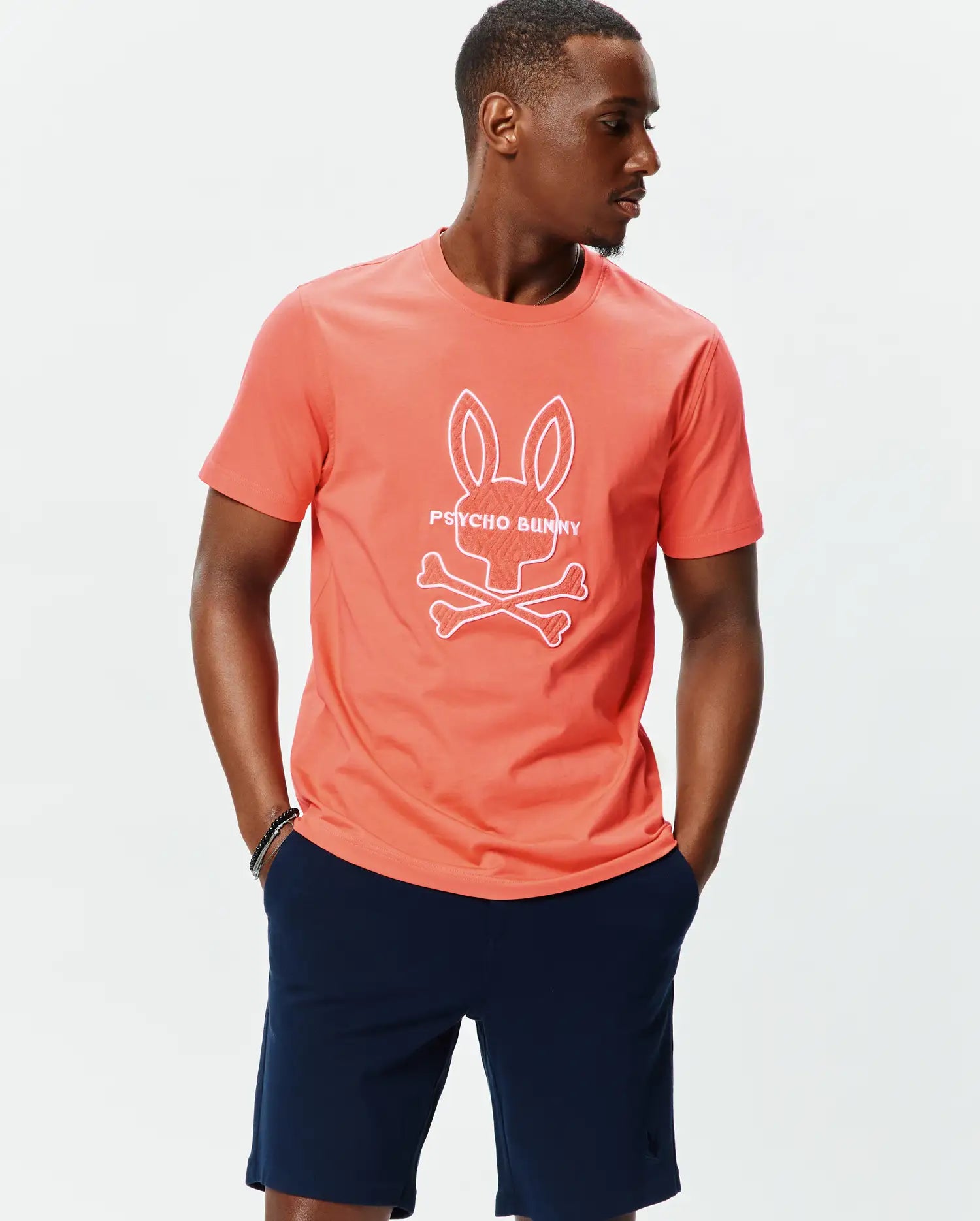 Mens Craig Graphic Tee, a classic fit coral jersey t-shirt featuring a large white outline Psycho Bunny skull and crossbones logo with text prominently on the center chest. (6)