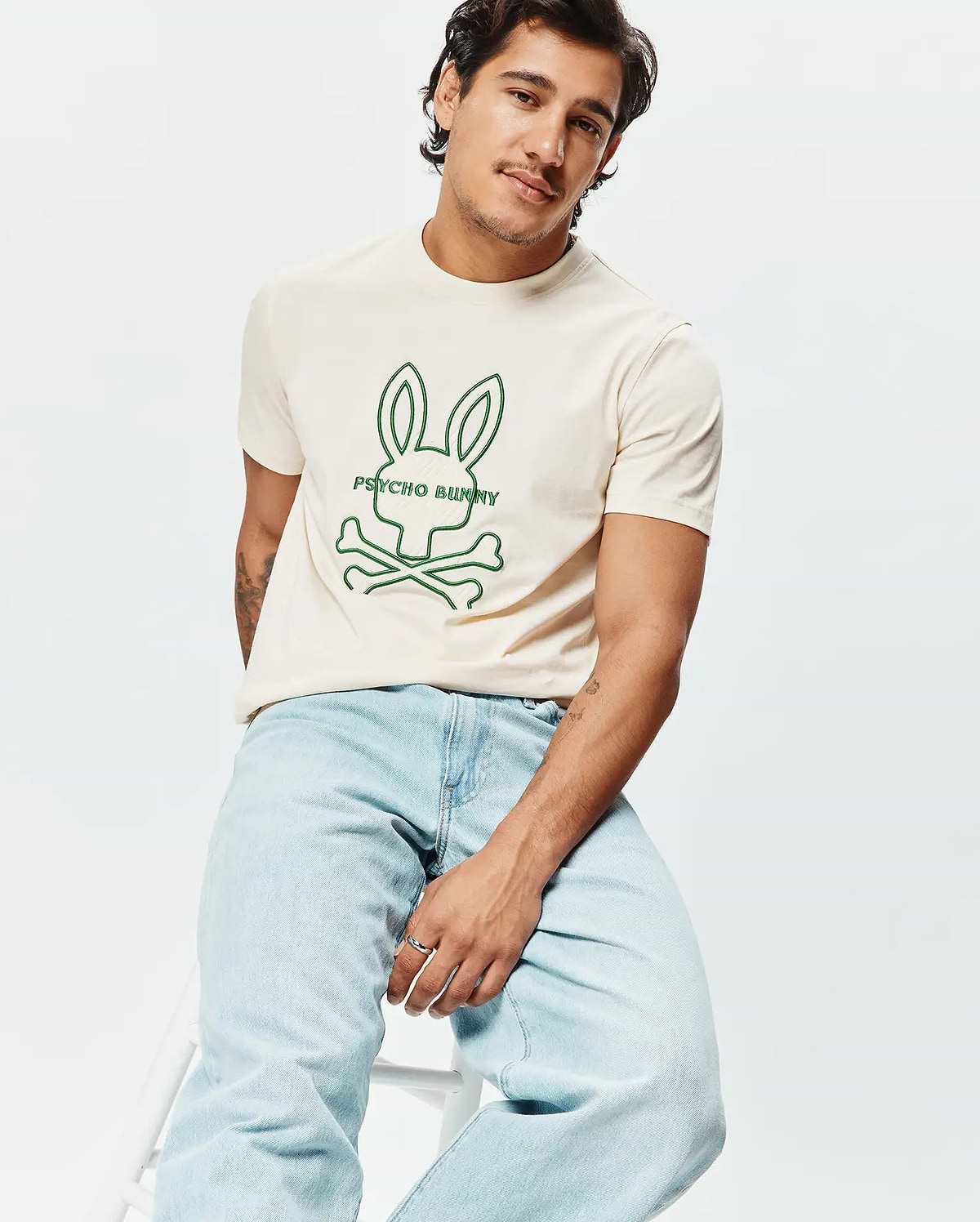 Mens Craig Graphic Tee, a classic fit cream jersey t-shirt with a prominent green outline graphic of the iconic Psycho Bunny skull and crossbones logo and text centered on the chest. (4)