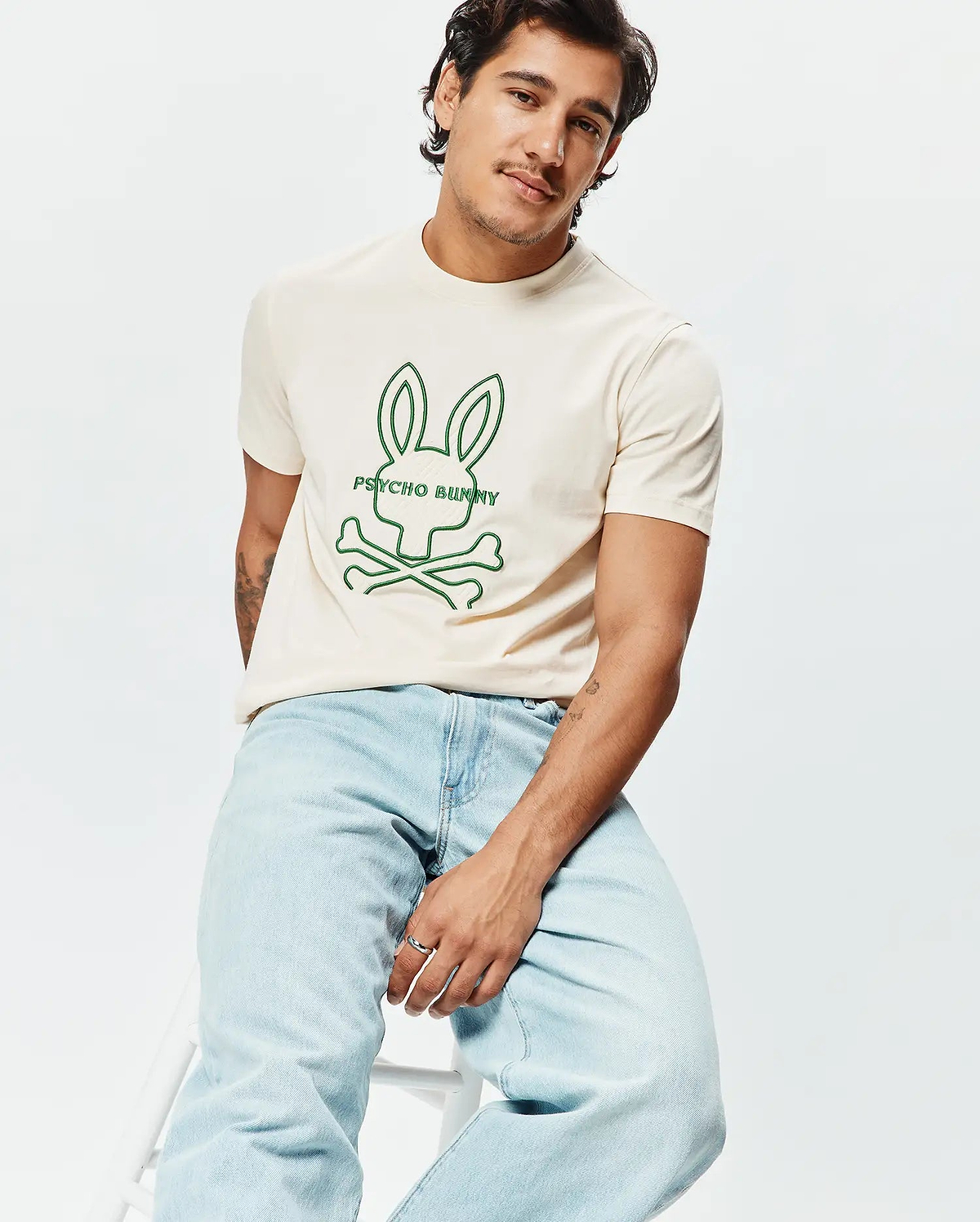 Mens Craig Graphic Tee, a classic fit cream jersey t-shirt with a prominent green outline graphic of the iconic Psycho Bunny skull and crossbones logo and text centered on the chest. (4)