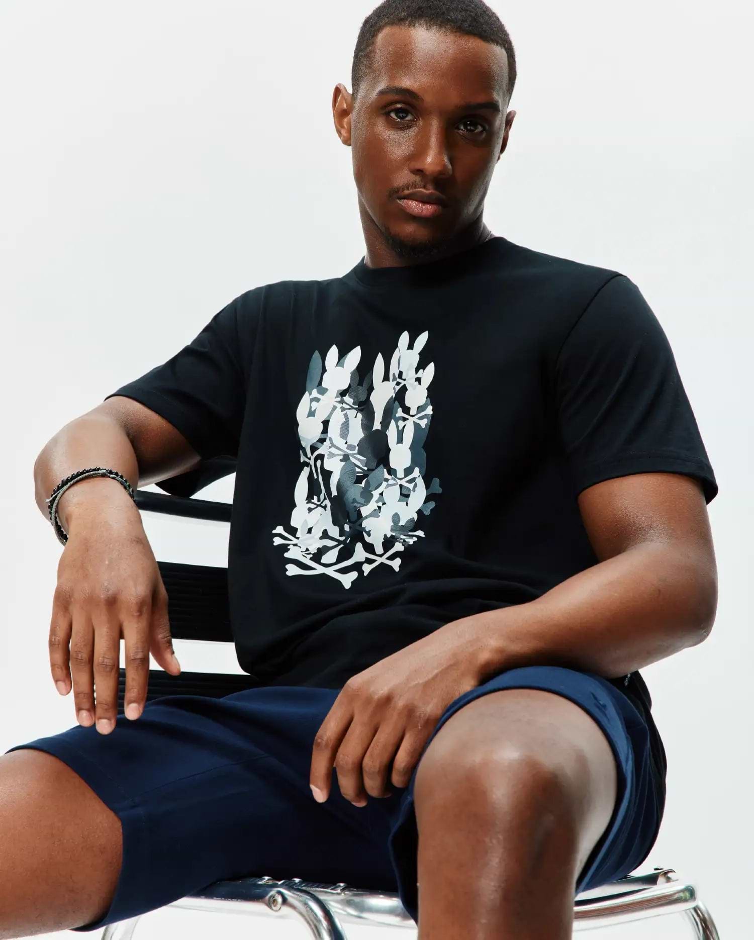 Mens Tanner Graphic Tee, a black jersey t-shirt with a large central graphic print of stacked white and grey Psycho Bunny skull logos and crossbones, classic fit.