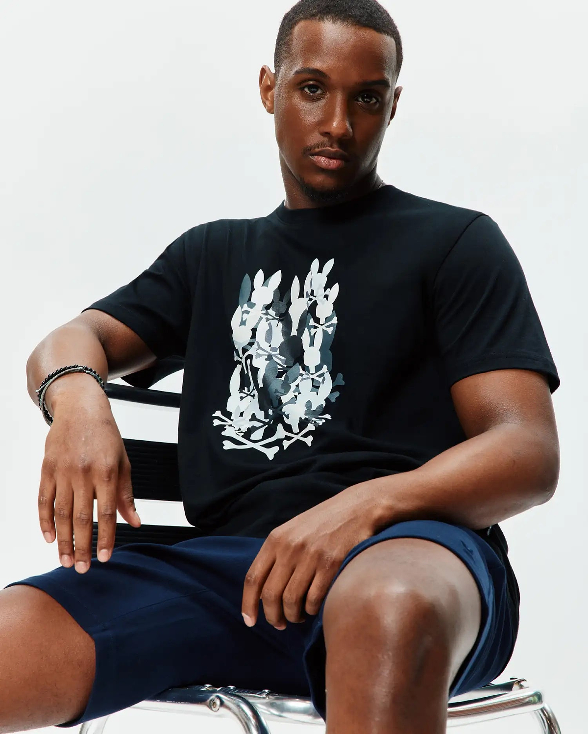 Mens Tanner Graphic Tee, a black jersey t-shirt with a large central graphic print of stacked white and grey Psycho Bunny skull logos and crossbones, classic fit.