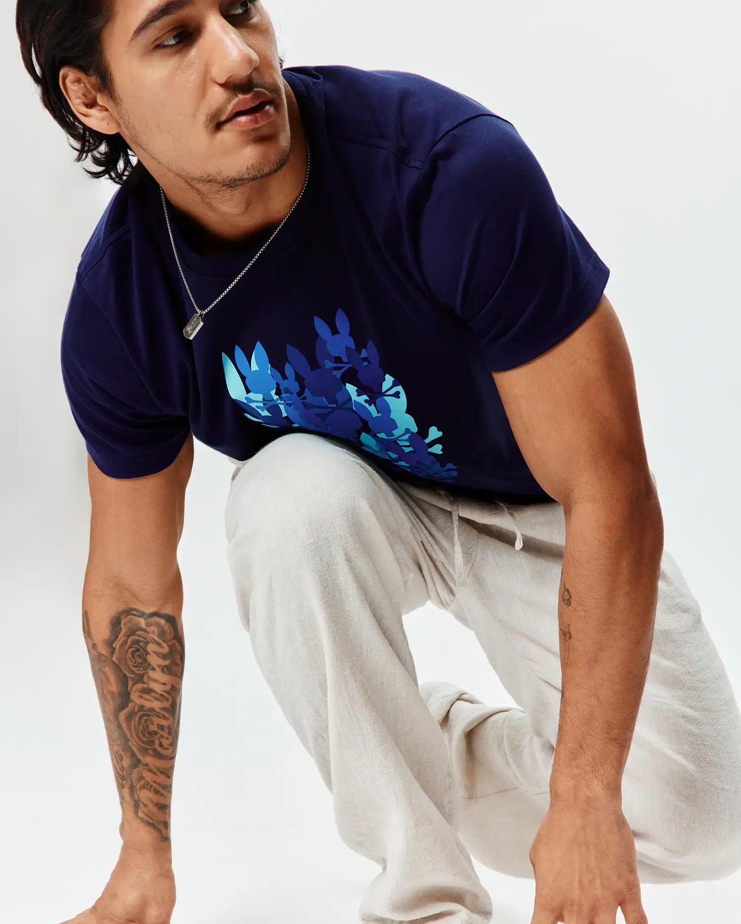 Mens Tanner Graphic Tee, a classic fit navy blue jersey t-shirt with a large chest graphic of multiple bunny skulls in teal, light blue, and darker blue. Paired with light beige textured drawstring pants.