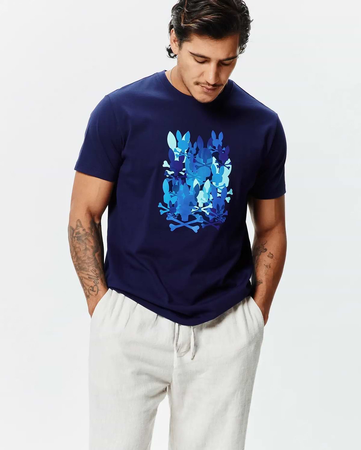 Mens Tanner Graphic Tee, a navy blue classic fit jersey t-shirt featuring a large central graphic of multiple Psycho Bunny skull and crossbones logos in various shades of blue.