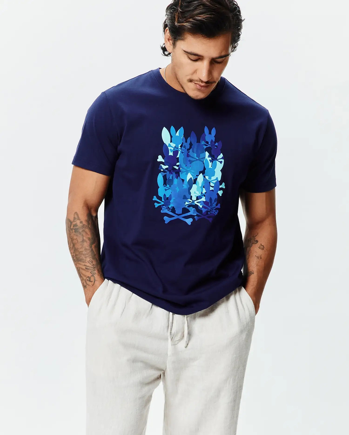 Mens Tanner Graphic Tee, a navy blue classic fit jersey t-shirt featuring a large central graphic of multiple Psycho Bunny skull and crossbones logos in various shades of blue.