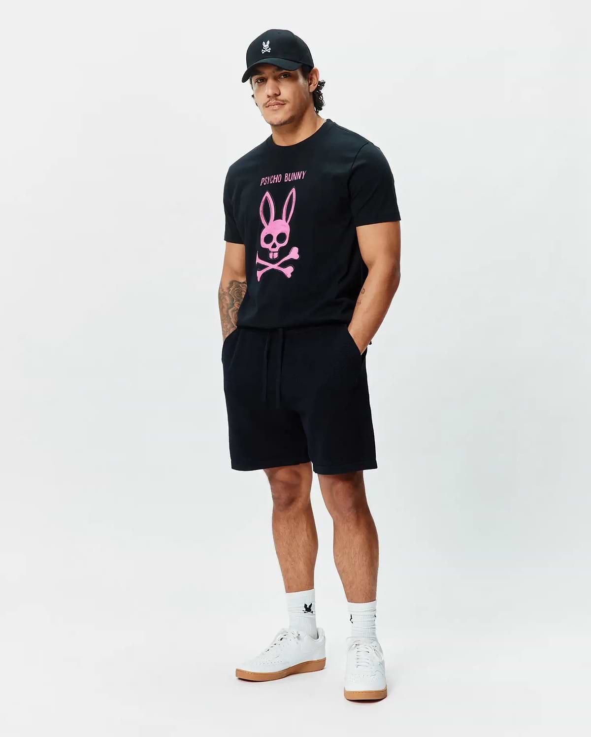 Mens Adler Graphic Tee, a classic fit black jersey t-shirt with a large pink Psycho Bunny skull and crossbones chest graphic. Black French terry shorts, a black cap, and white socks with bunny skull logos. (5)