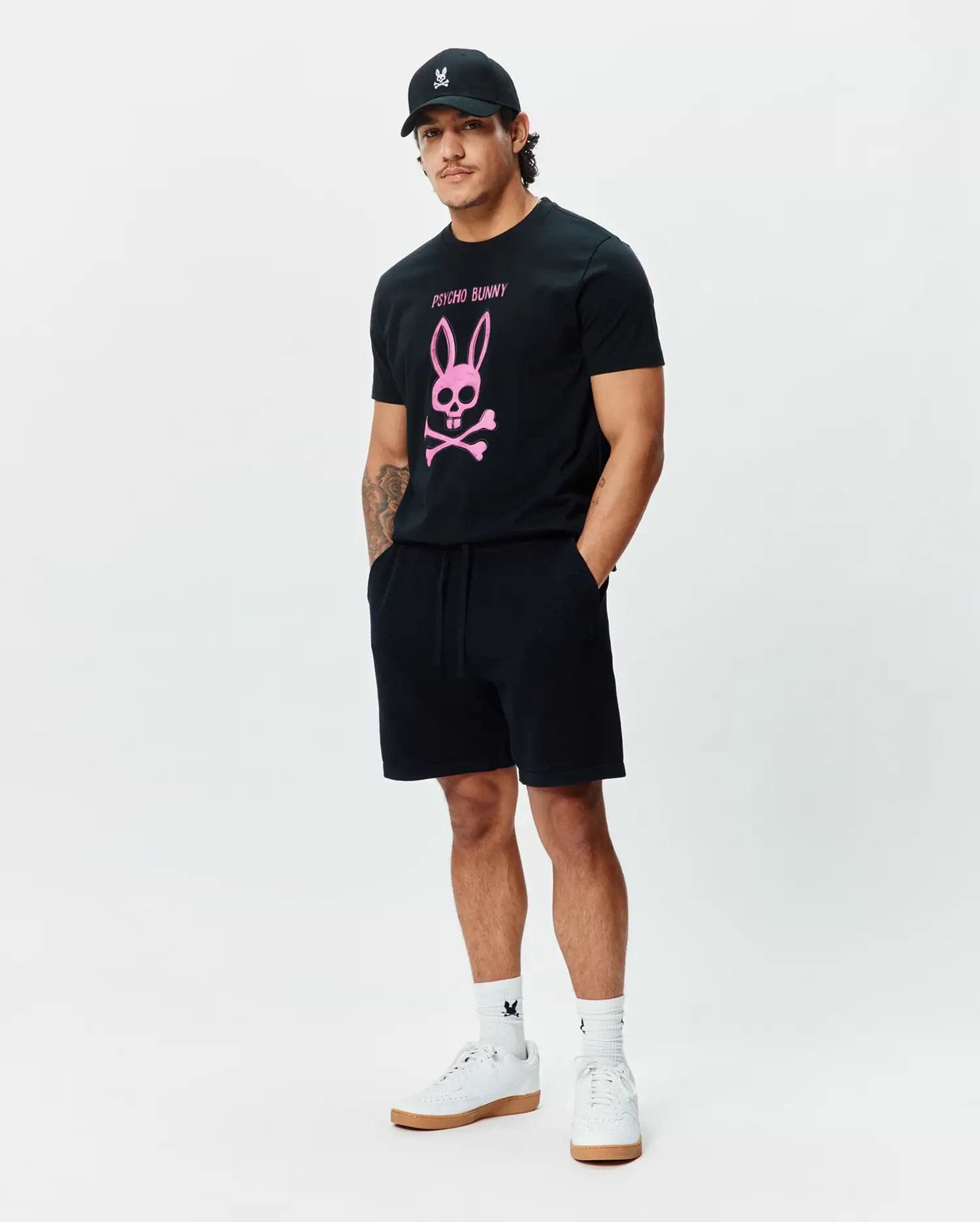 Mens Adler Graphic Tee, a classic fit black jersey t-shirt with a large pink Psycho Bunny skull and crossbones chest graphic. Black French terry shorts, a black cap, and white socks with bunny skull logos. (5)