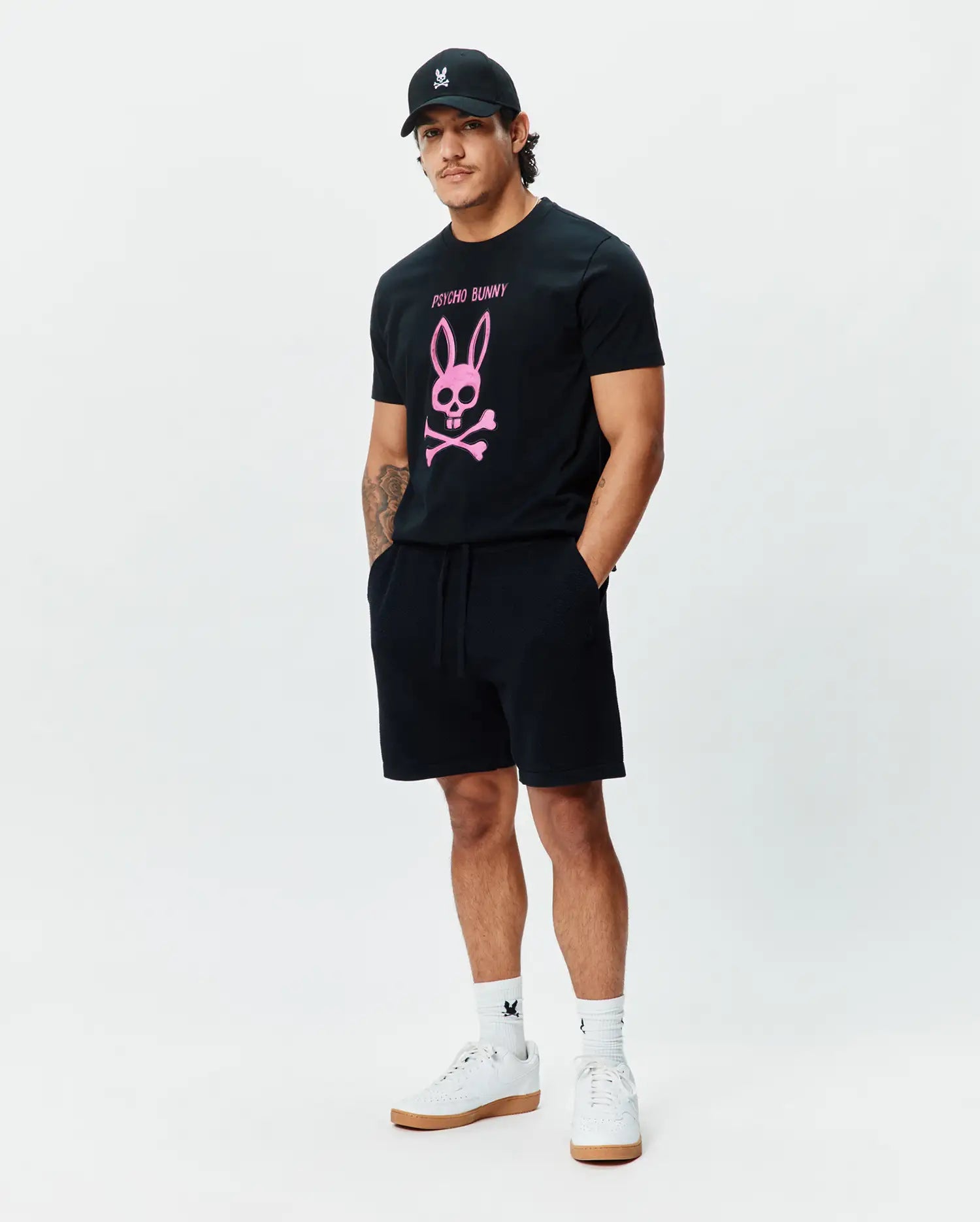 Mens Adler Graphic Tee, a classic fit black jersey t-shirt with a large pink Psycho Bunny skull and crossbones chest graphic. Black French terry shorts, a black cap, and white socks with bunny skull logos. (5)