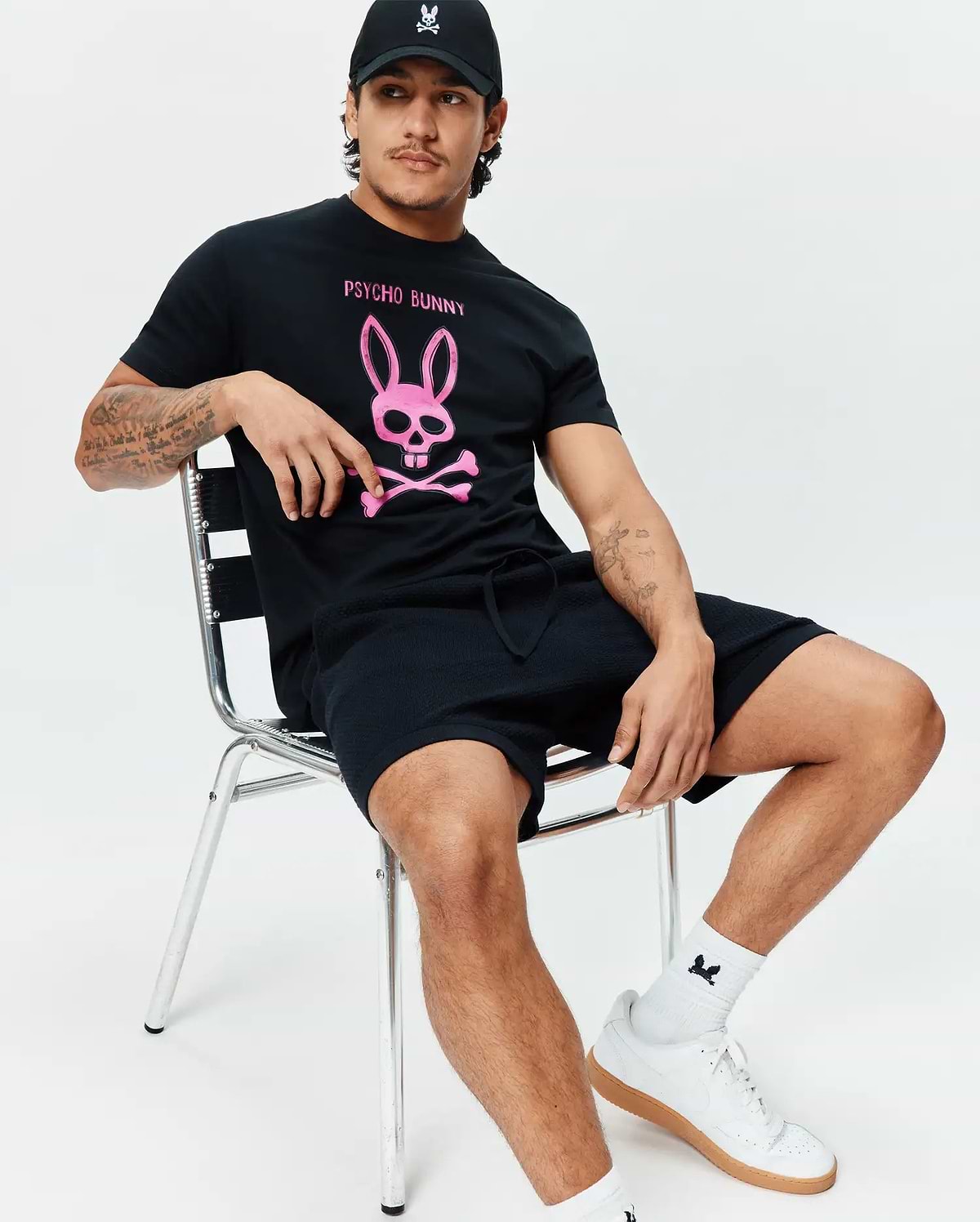 Black Mens Adler Graphic Tee featuring a bold pink Psycho Bunny skull graphic, paired with black textured drawstring shorts, a black cap with a white embroidered bunny logo, and white crew socks with a small black bunny. (2)