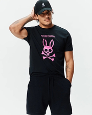 Black jersey Mens Adler Graphic Tee with a large pink Psycho Bunny skull and crossbones graphic on the chest, paired with black textured shorts and a black cap with a white bunny logo. (4)