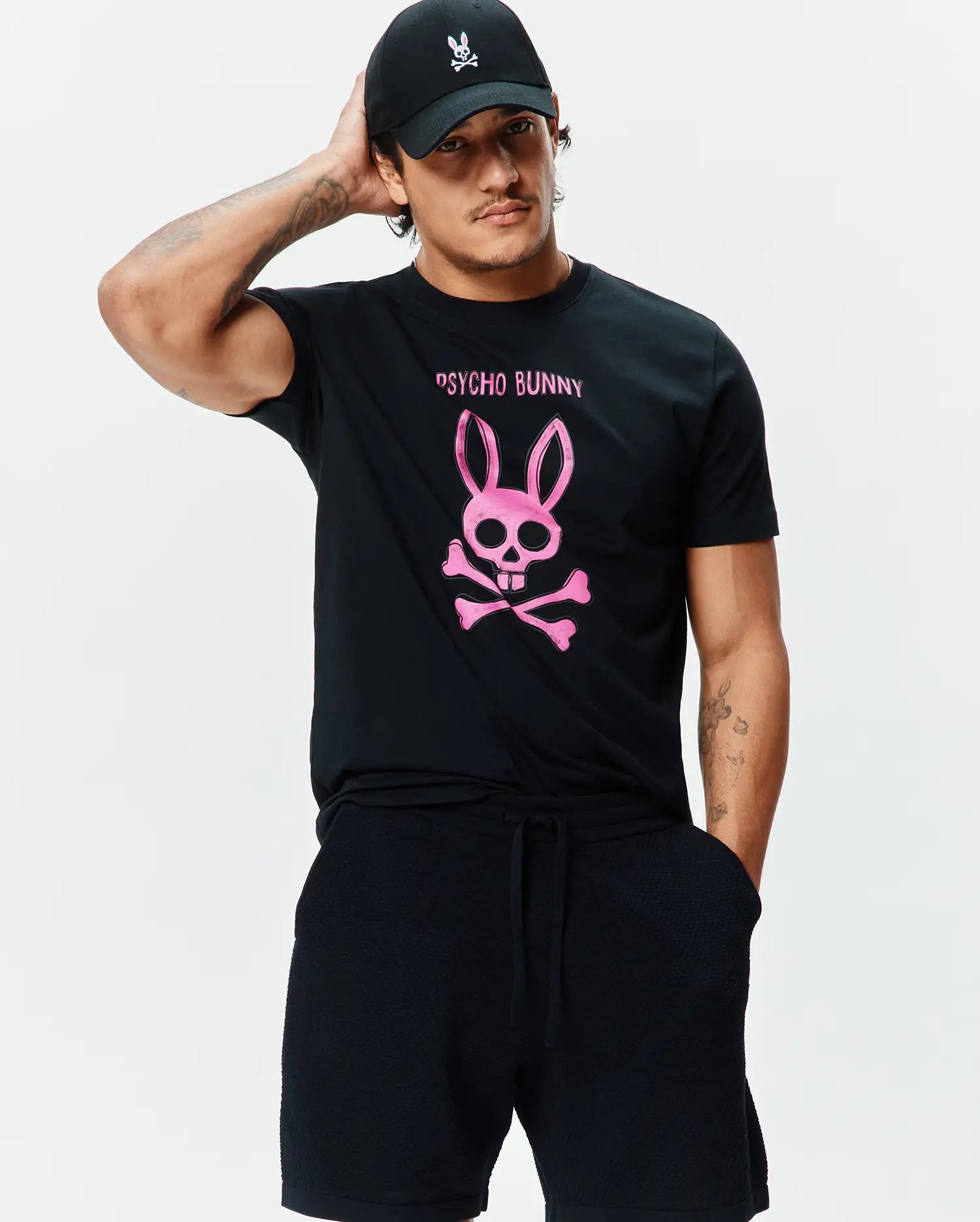 Black jersey Mens Adler Graphic Tee with a large pink Psycho Bunny skull and crossbones graphic on the chest, paired with black textured shorts and a black cap with a white bunny logo. (4)