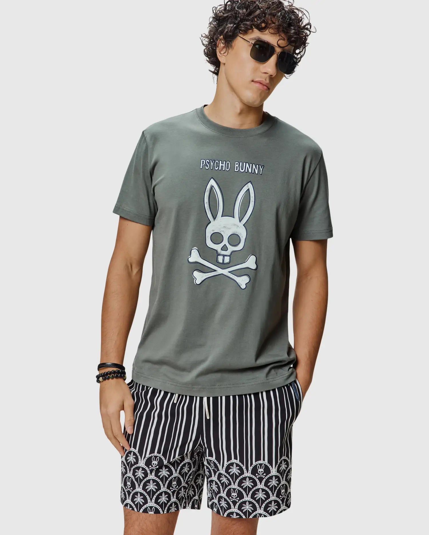 Mens Adler Graphic Tee, an olive grey classic fit jersey knit t-shirt featuring a large white Psycho Bunny skull and crossbones graphic with dark blue outline and PSYCHO BUNNY text on the chest.