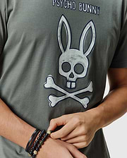 Mens Adler Graphic Tee in dark olive green, featuring a large distressed white and blue Psycho Bunny skull and crossbones graphic on the chest, made from soft jersey knit.