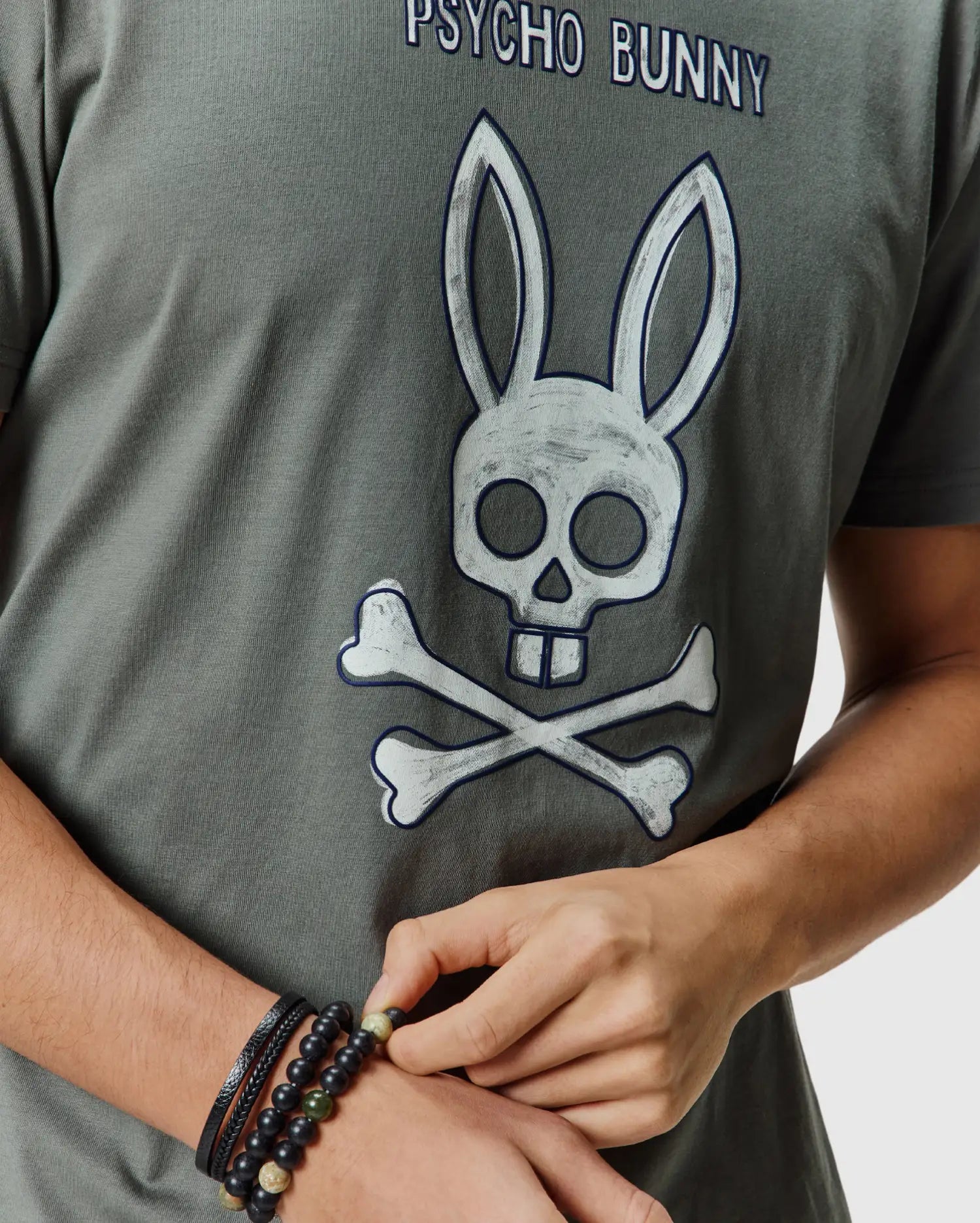 Mens Adler Graphic Tee in dark olive green, featuring a large distressed white and blue Psycho Bunny skull and crossbones graphic on the chest, made from soft jersey knit.