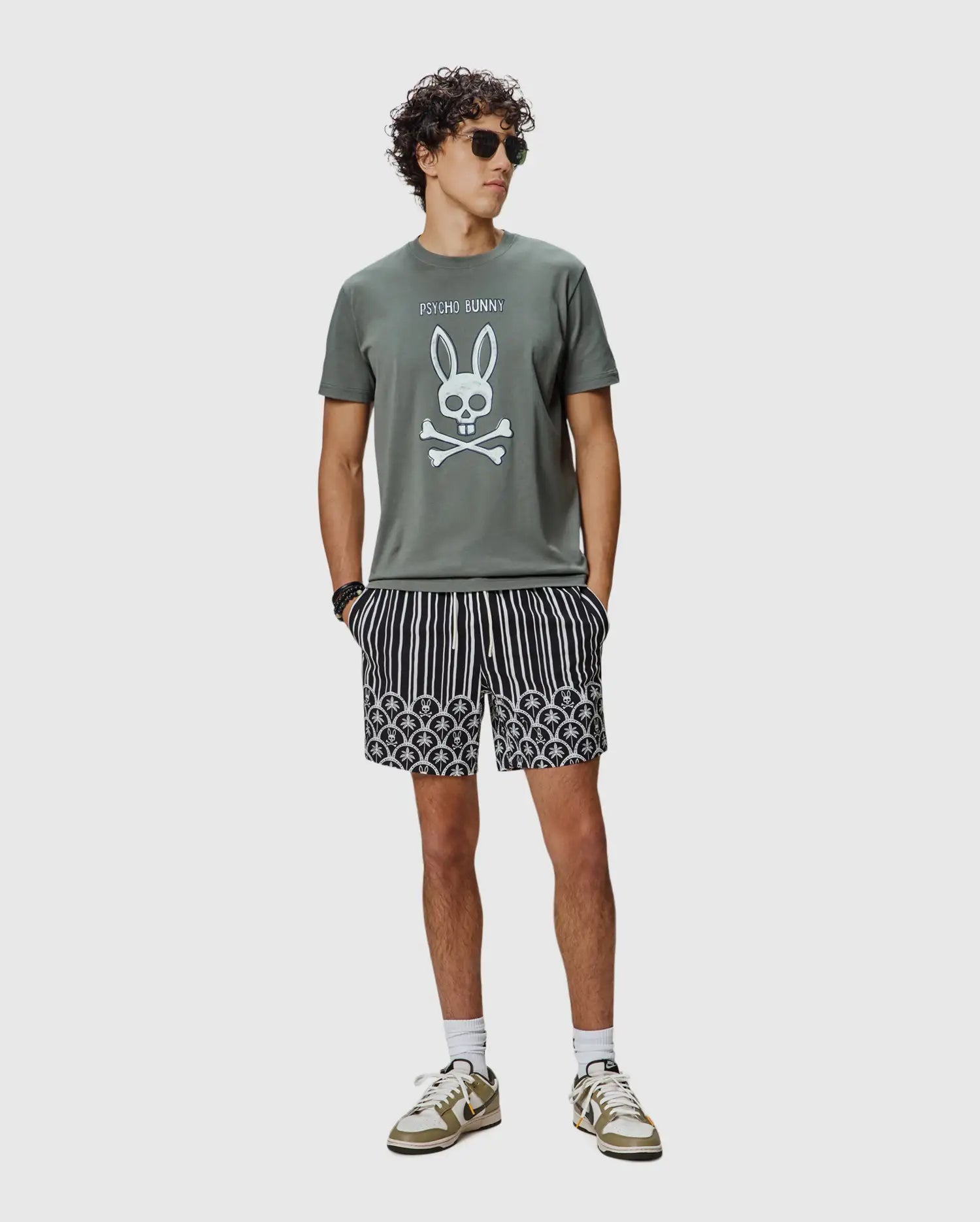 Mens Adler Graphic Tee in olive green jersey with a large white Psycho Bunny skull and crossbones graphic, paired with black and white striped shorts featuring a lower scalloped pattern of small bunny skull logos.