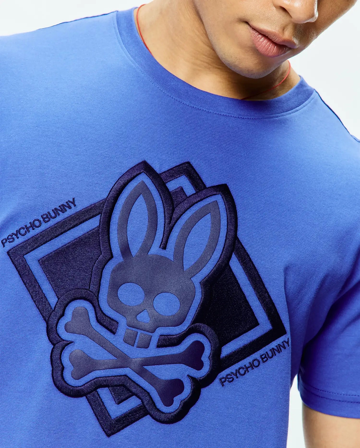 Mens Tobias Graphic Tee, a vibrant blue jersey t-shirt with a large, central raised Psycho Bunny skull logo in layered dark blue and black, set in a diamond graphic with diagonal Psycho Bunny text. (3)