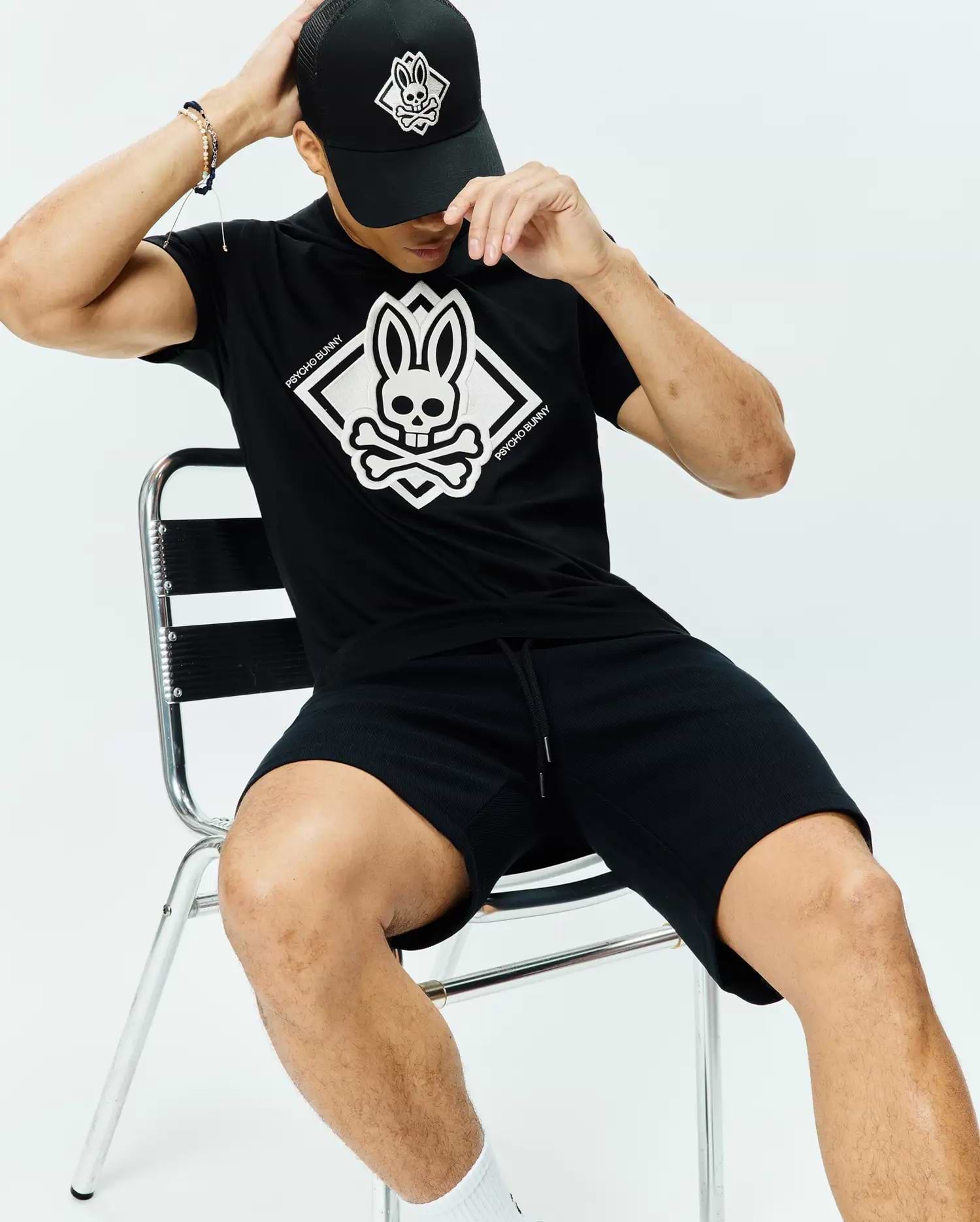 Black Mens Tobias Graphic Tee, classic jersey fit, with a large white Psycho Bunny skull and crossbones diamond chest graphic. Paired with black textured drawstring shorts and a black mesh cap with white logo.