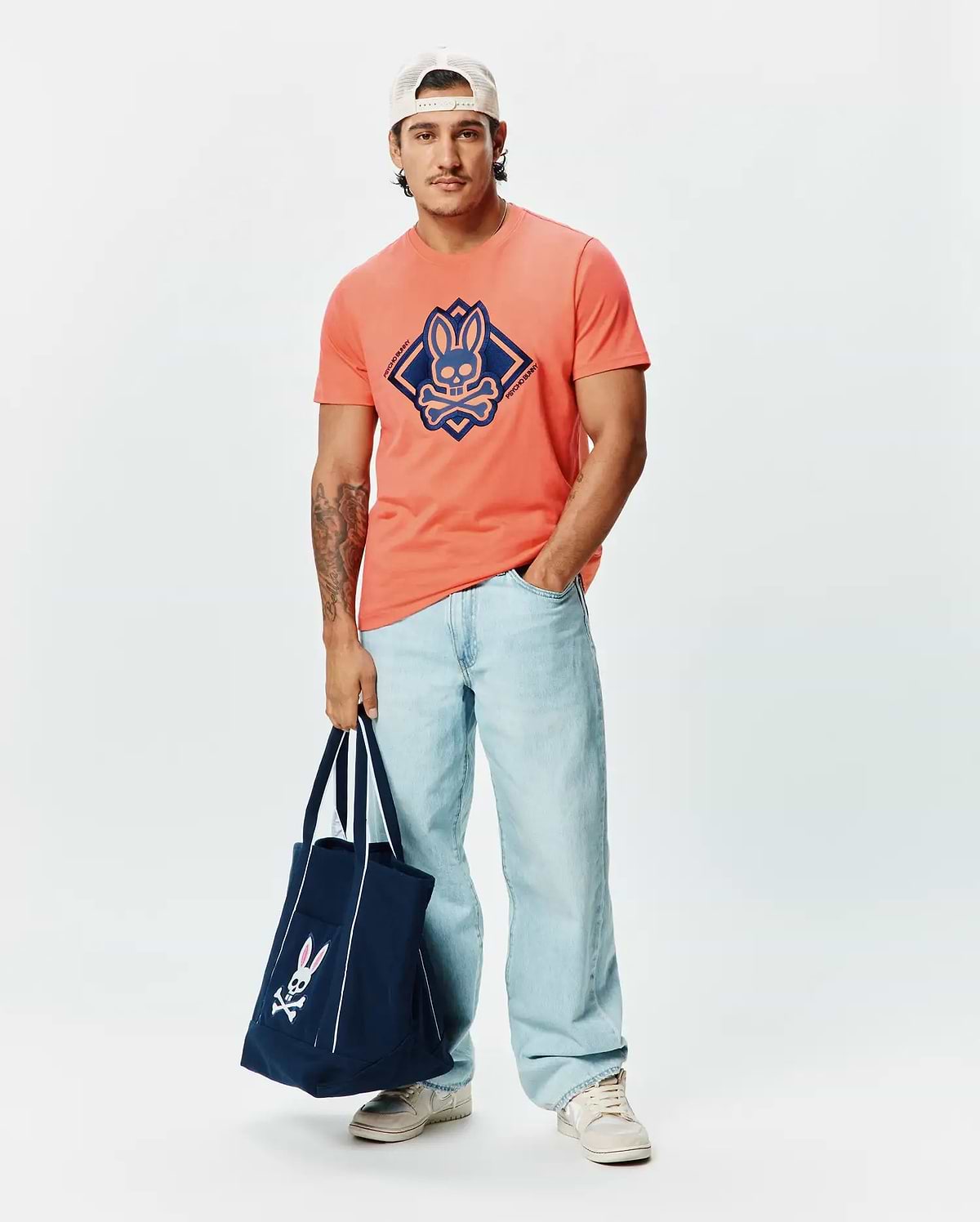 Mens Tobias Graphic Tee, a classic fit coral jersey t-shirt with a large, centered navy blue Psycho Bunny skull logo within a diamond graphic, accented by brand text.