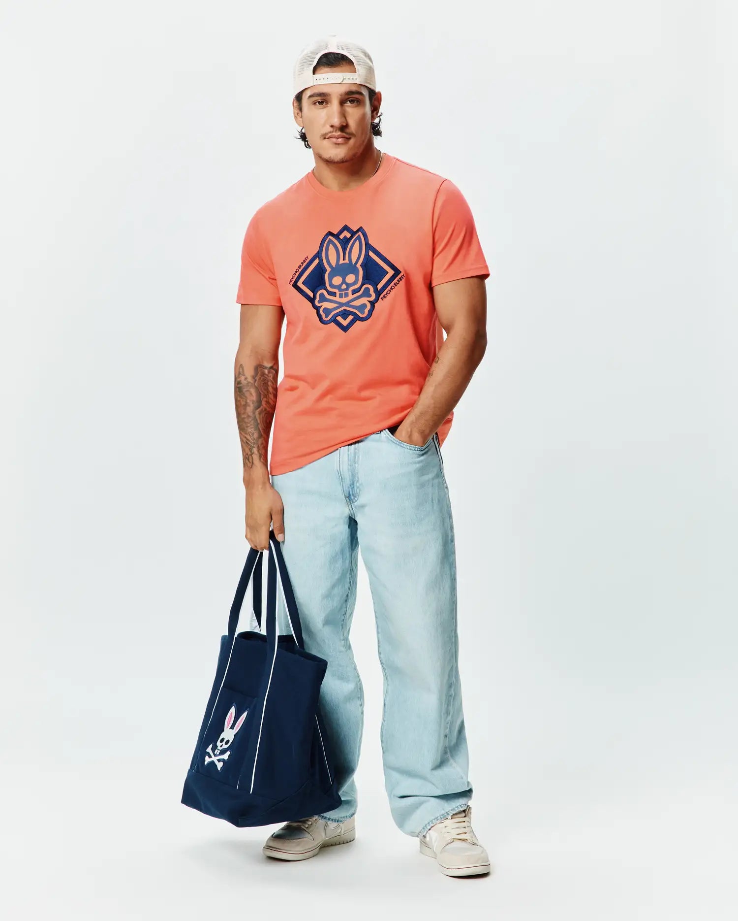 Mens Tobias Graphic Tee, a classic fit coral jersey t-shirt with a large, centered navy blue Psycho Bunny skull logo within a diamond graphic, accented by brand text.