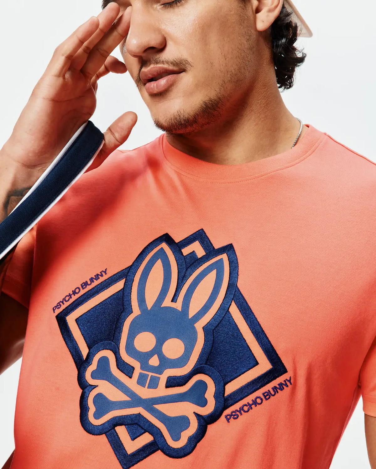 Mens Tobias Graphic Tee, a classic fit coral jersey t-shirt featuring a large central navy and light blue Psycho Bunny skull and crossbones graphic, outlined in coral, with embroidered Psycho Bunny text.