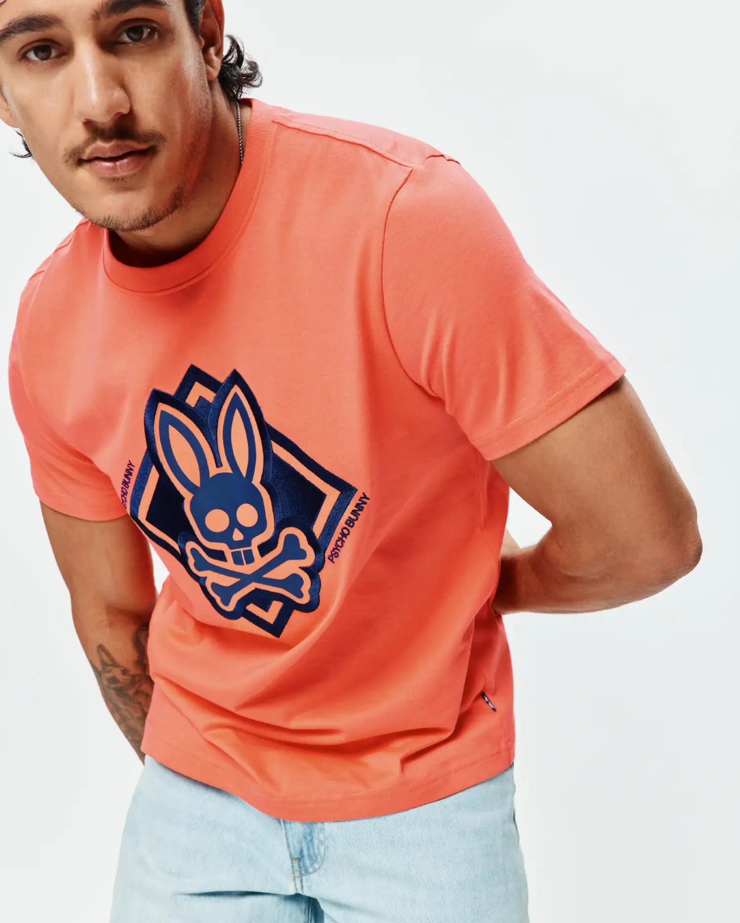 Mens Tobias Graphic Tee in coral, with a large navy blue Psycho Bunny skull and crossbones graphic on the chest, framed by layered diamonds and Psycho Bunny text, classic fit, smooth jersey.