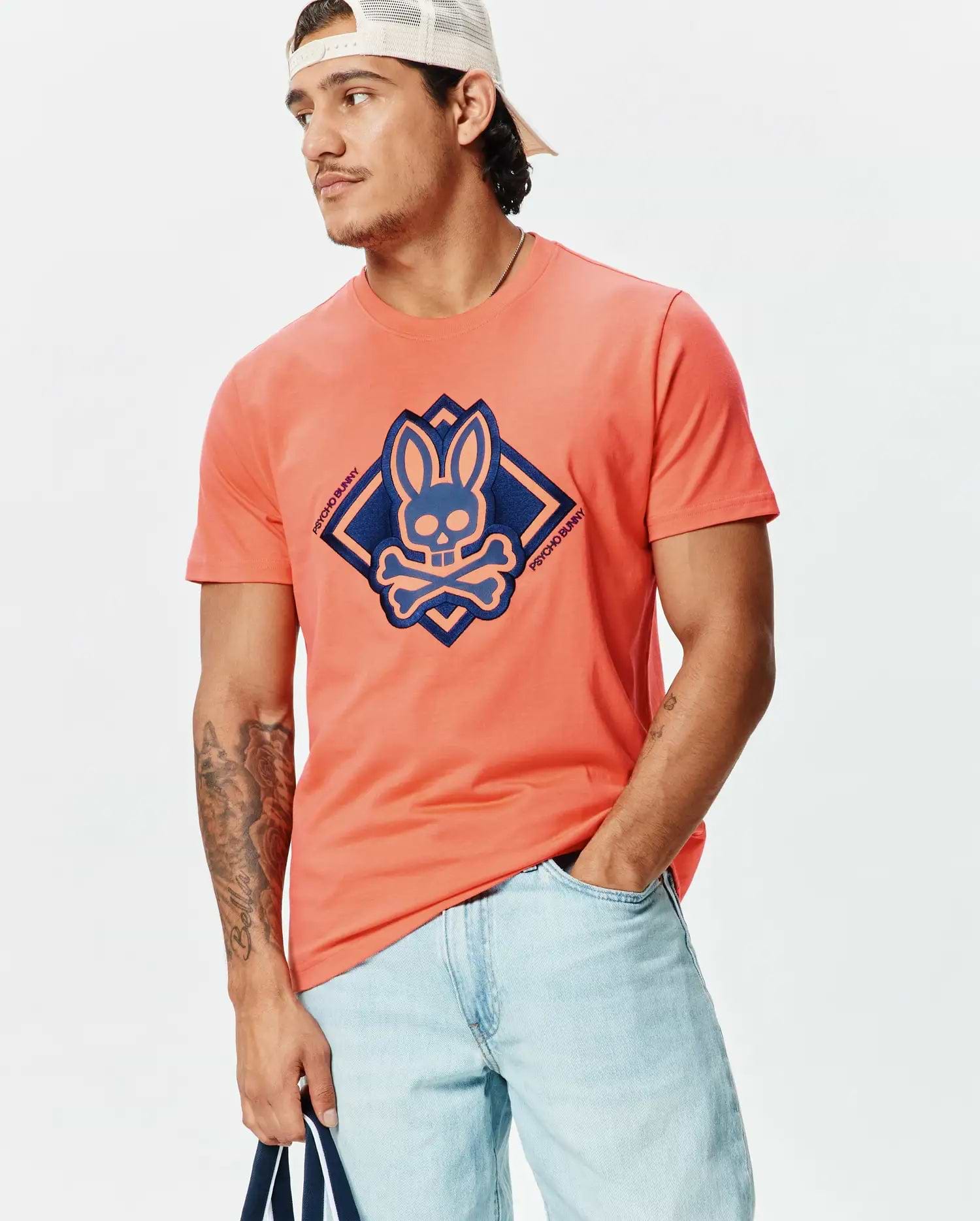 Mens Tobias Graphic Tee, a coral jersey knit t-shirt with a classic fit, featuring a large central dark blue and light blue Psycho Bunny skull and crossbones logo within a diamond graphic on the chest.
