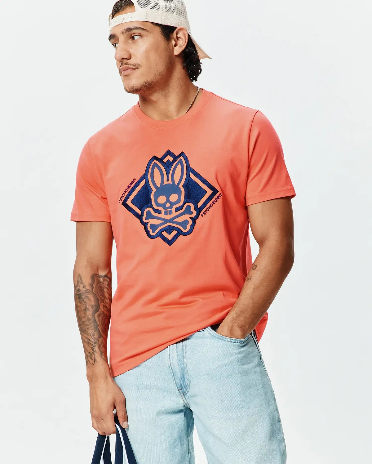 Mens Tobias Graphic Tee, a coral jersey knit t-shirt with a classic fit, featuring a large central dark blue and light blue Psycho Bunny skull and crossbones logo within a diamond graphic on the chest.