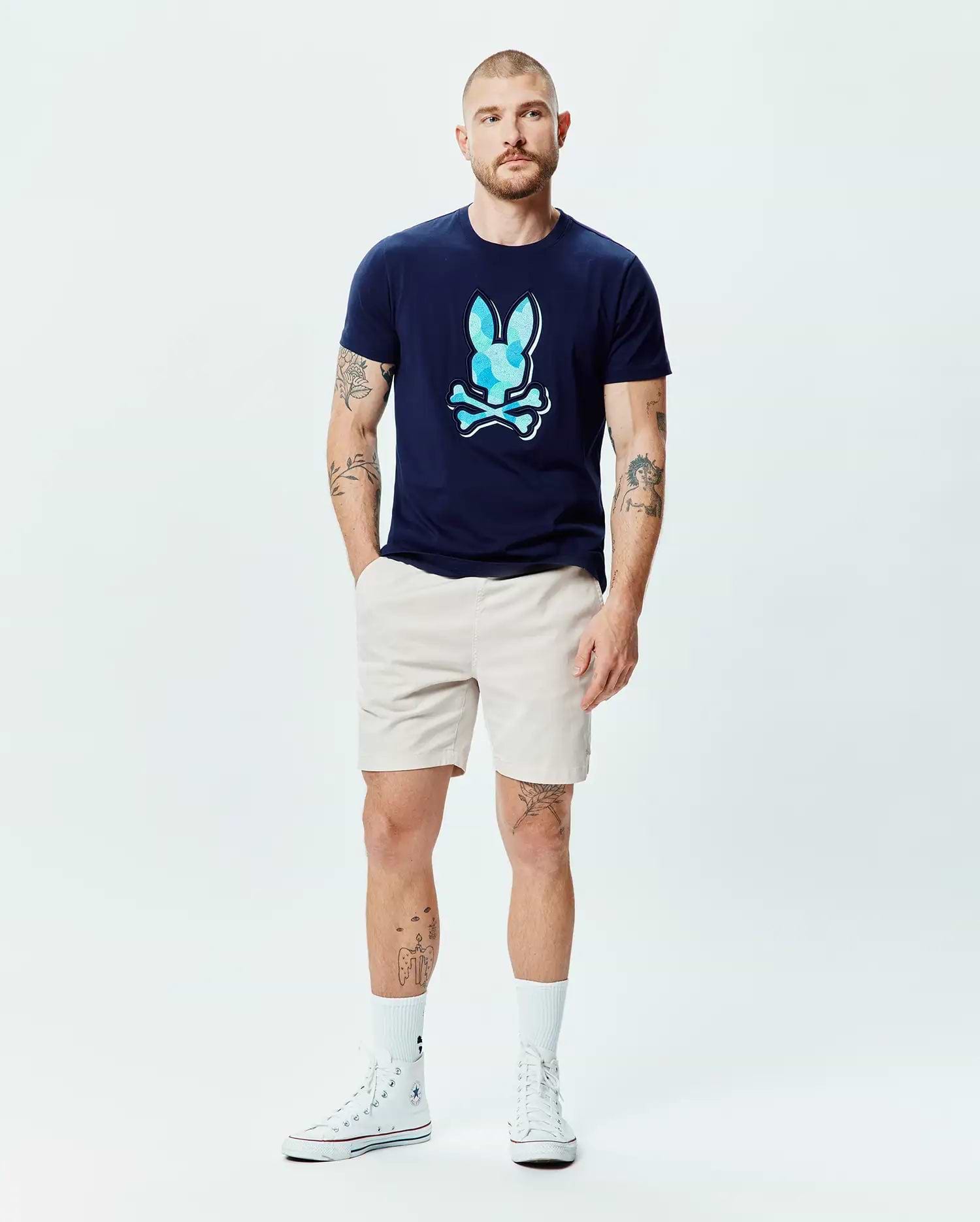 Mens Ethan Graphic Tee in navy blue jersey, featuring a large, classic fit, light blue faceted Psycho Bunny skull logo on the chest, paired with classic beige shorts. (6)