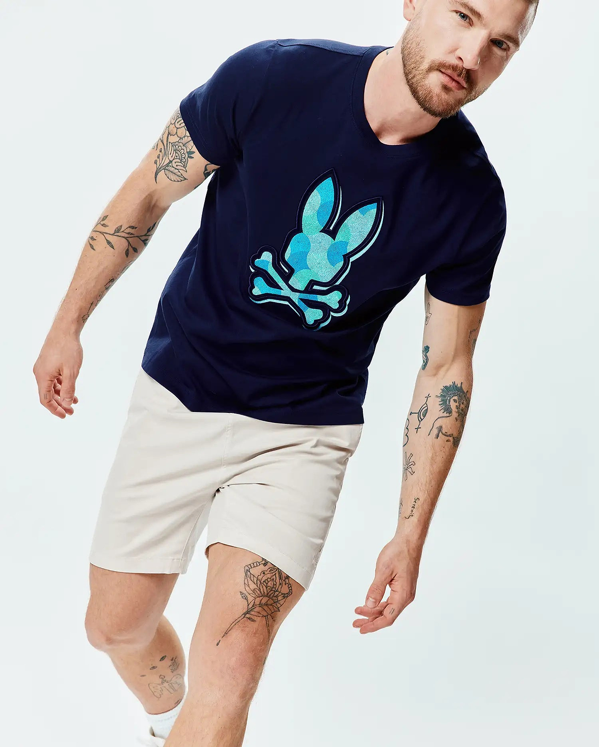 Mens Ethan Graphic Tee, navy blue classic fit jersey t-shirt with a large central blue and teal geometric Psycho Bunny skull logo, paired with light beige classic fit shorts. (5)