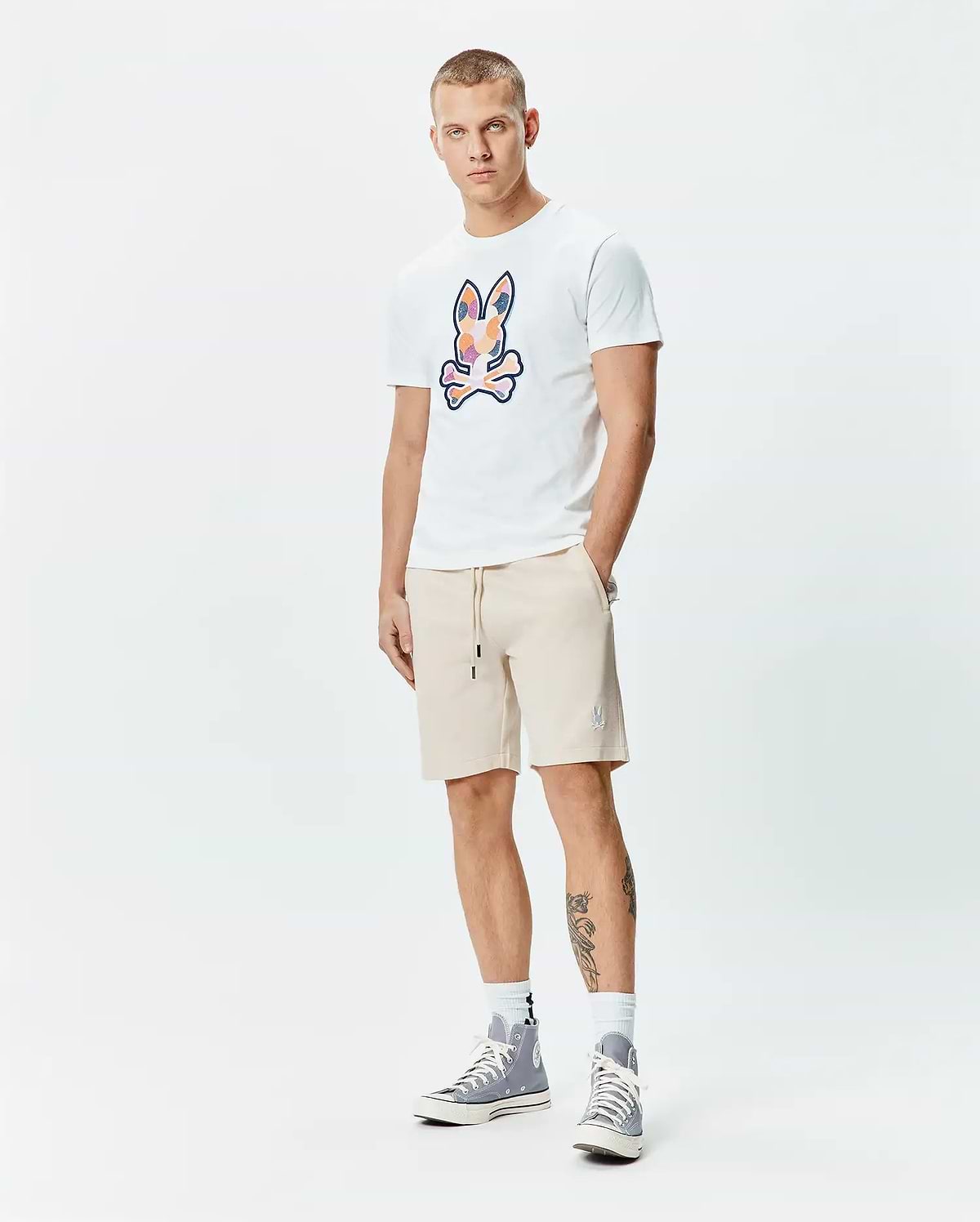 Mens Ethan Graphic Tee, a white jersey t-shirt with a large, colorful abstract Psycho Bunny skull logo on the chest, paired with beige french terry shorts featuring a small embroidered bunny logo on the left thigh and a drawstring waist. (5)