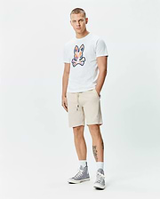 Mens Ethan Graphic Tee, a white jersey t-shirt with a large, colorful abstract Psycho Bunny skull logo on the chest, paired with beige french terry shorts featuring a small embroidered bunny logo on the left thigh and a drawstring waist. (5)