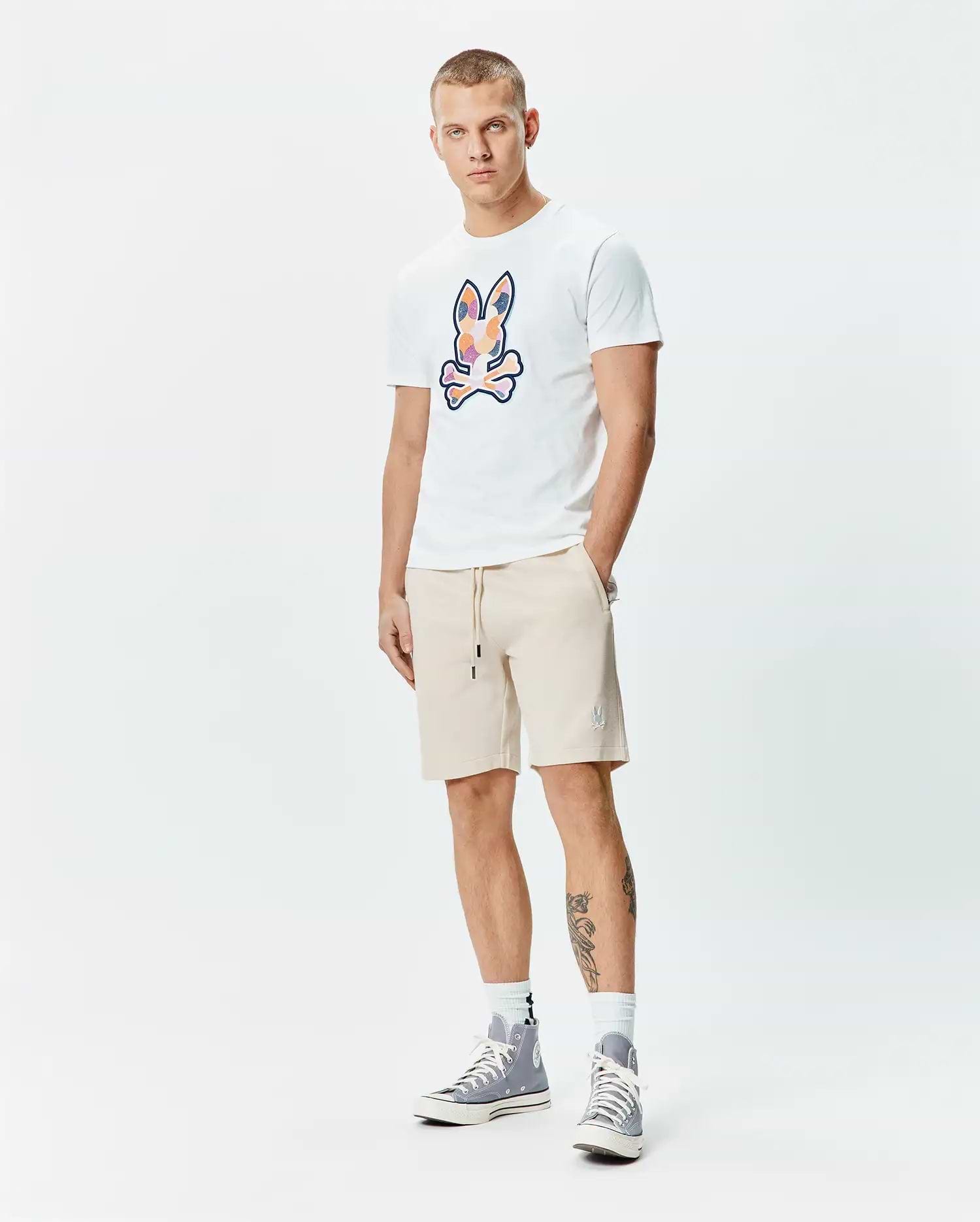Mens Ethan Graphic Tee, a white jersey t-shirt with a large, colorful abstract Psycho Bunny skull logo on the chest, paired with beige french terry shorts featuring a small embroidered bunny logo on the left thigh and a drawstring waist. (5)