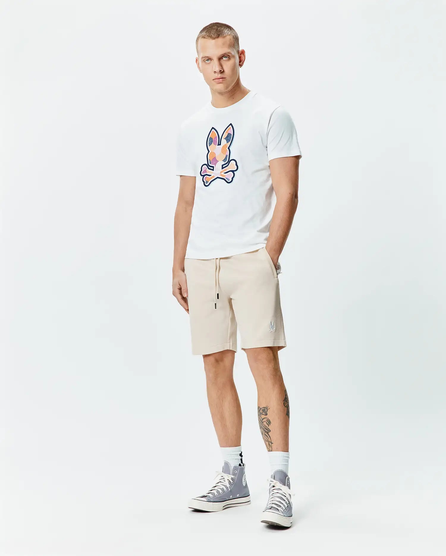 Mens Ethan Graphic Tee, a white jersey t-shirt with a large, colorful abstract Psycho Bunny skull logo on the chest, paired with beige french terry shorts featuring a small embroidered bunny logo on the left thigh and a drawstring waist. (5)