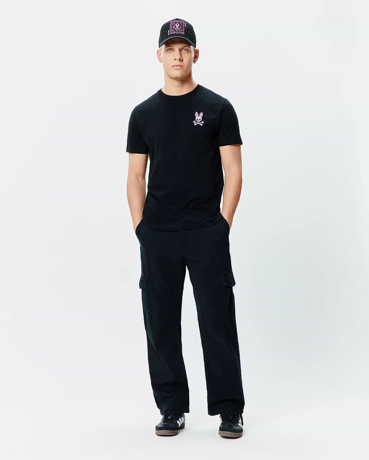Mens Frank Back Graphic Tee in black jersey, featuring a small pink embroidered Psycho Bunny skull logo on the left chest, paired with black relaxed-fit cargo pants and a black cap with a vibrant pink patterned front.