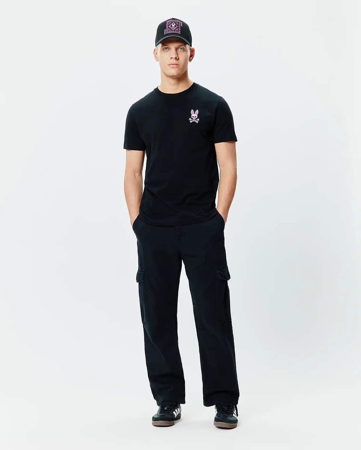 Mens Frank Back Graphic Tee in black jersey, featuring a small pink embroidered Psycho Bunny skull logo on the left chest, paired with black relaxed-fit cargo pants and a black cap with a vibrant pink patterned front.