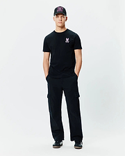Mens Frank Back Graphic Tee in black jersey, featuring a small pink embroidered Psycho Bunny skull logo on the left chest, paired with black relaxed-fit cargo pants and a black cap with a vibrant pink patterned front.