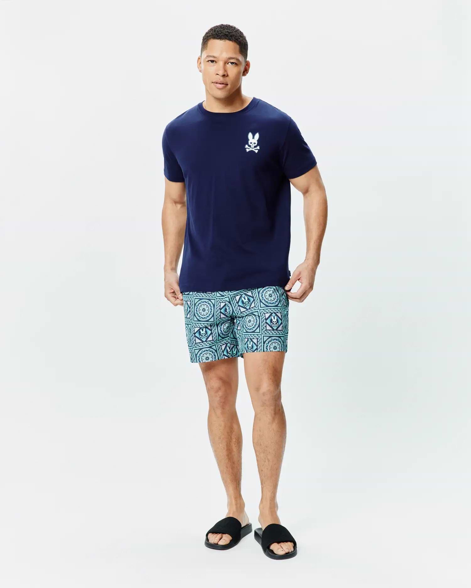 Navy blue classic fit jersey Mens Frank Back Graphic Tee with a small white embroidered Psycho Bunny skull logo on the chest, paired with teal geometric patterned shorts featuring repeated bunny skull motifs.