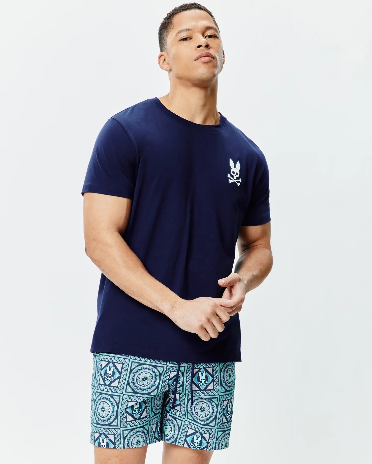 Navy blue classic fit jersey tee, Mens Frank Back Graphic Tee, with a white embroidered Psycho Bunny skull logo on left chest, paired with teal geometric shorts with integrated bunny skulls.