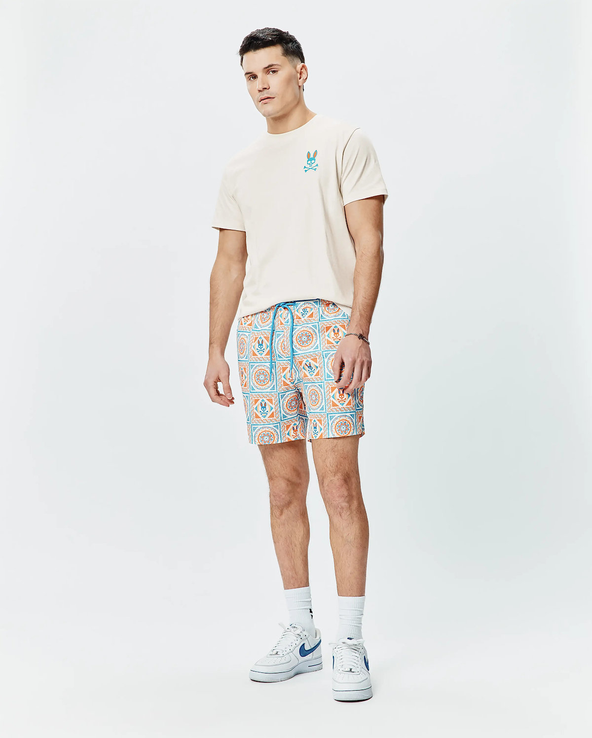 Mens Frank Back Graphic Tee, cream jersey with small blue/orange Psycho Bunny chest logo, and relaxed shorts featuring an orange/blue tiled pattern with bunny skull logos and a blue drawstring.
