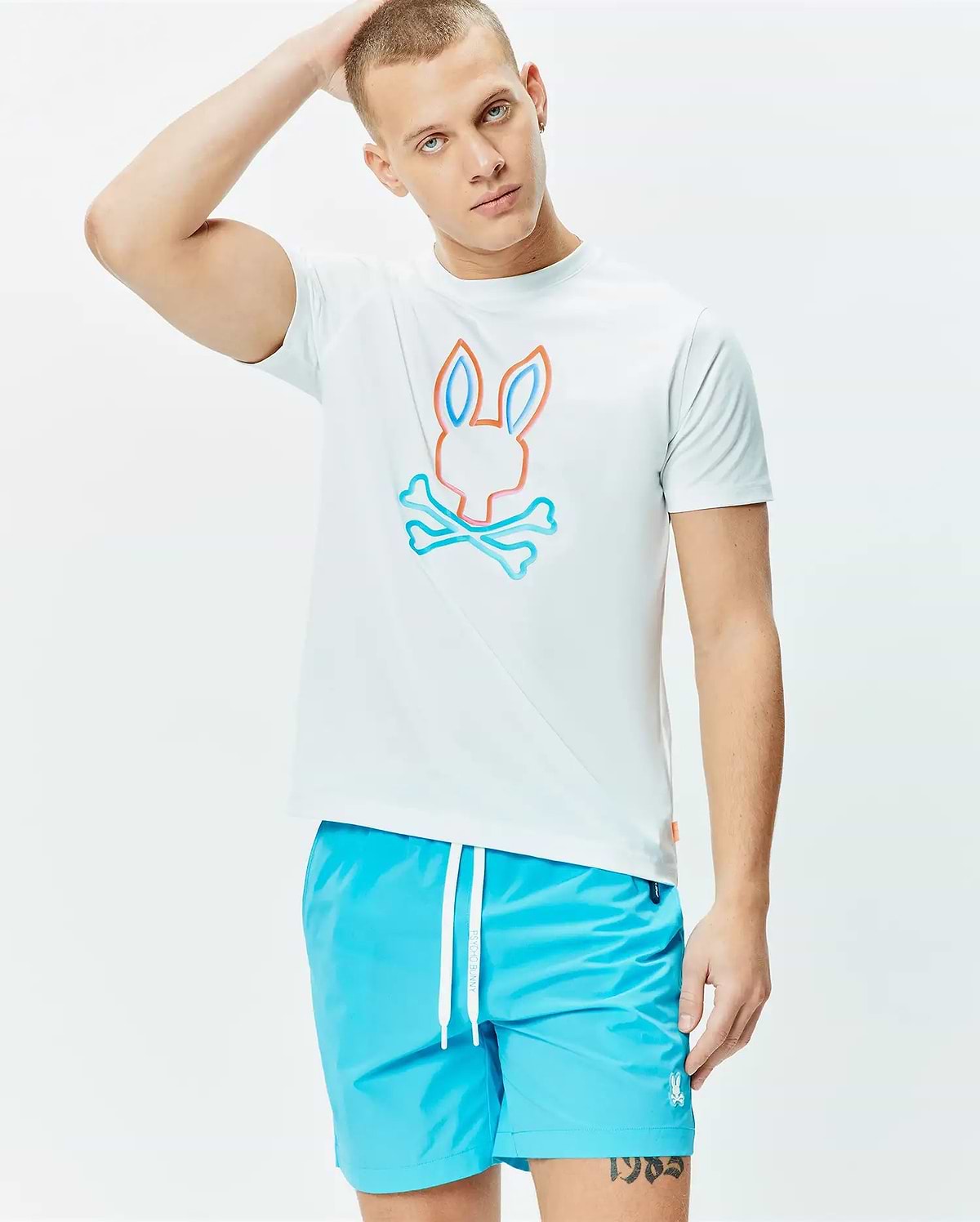 Mens Dayton Graphic Tee, a white classic fit jersey t-shirt featuring a large central Psycho Bunny skull logo outlined in vibrant orange and light blue, paired with bright blue swim shorts with a white drawstring and small embroidered bunny logo. (2)