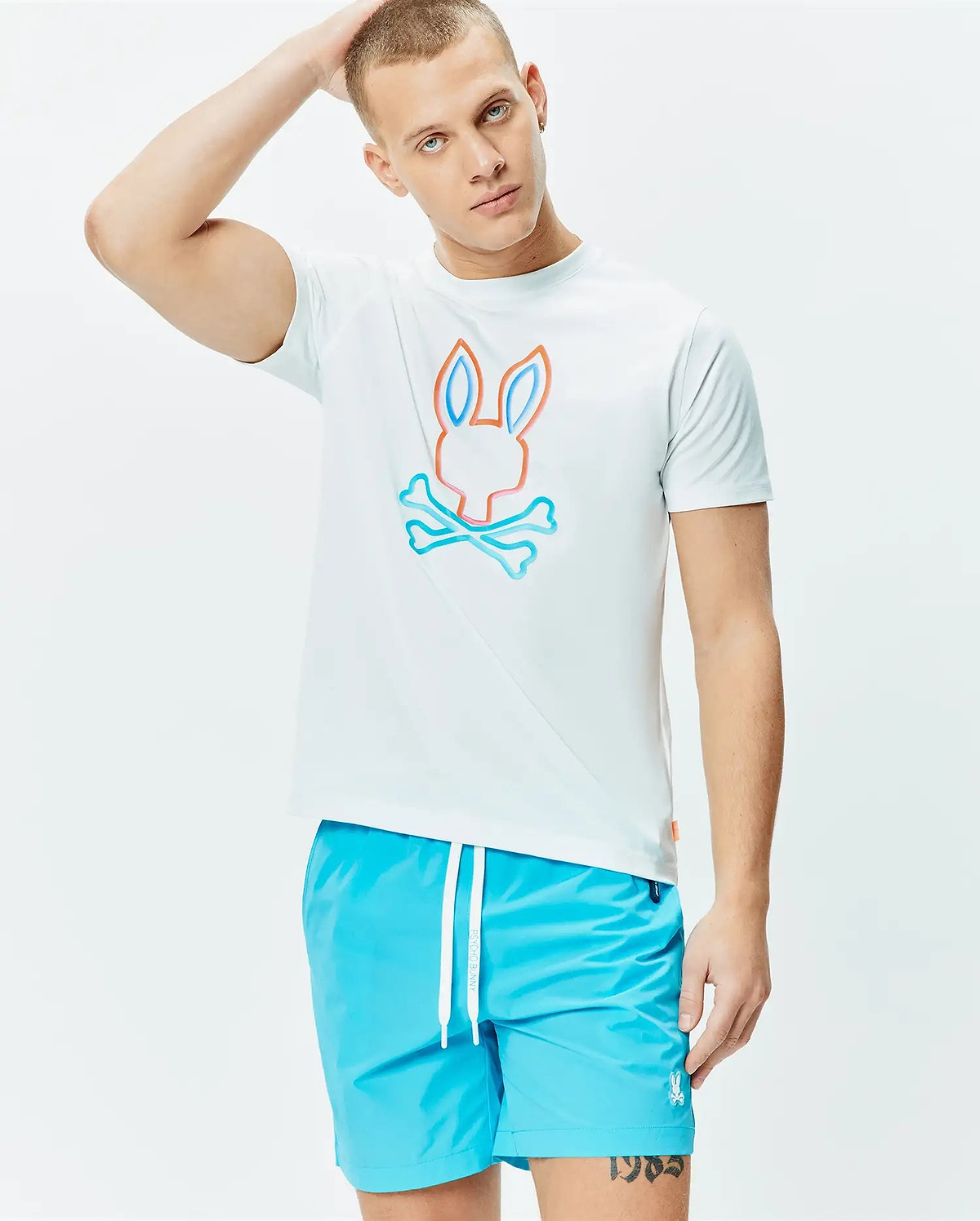 Mens Dayton Graphic Tee, a white classic fit jersey t-shirt featuring a large central Psycho Bunny skull logo outlined in vibrant orange and light blue, paired with bright blue swim shorts with a white drawstring and small embroidered bunny logo. (2)