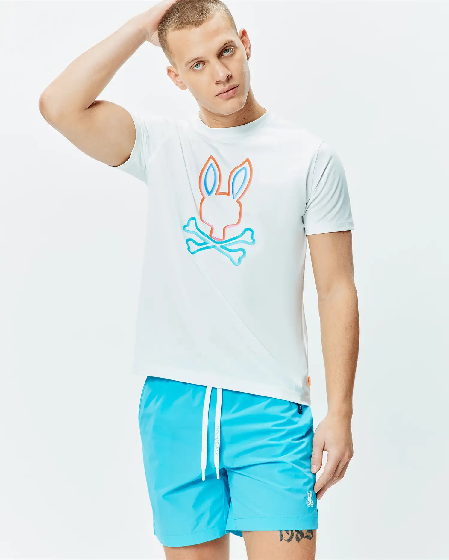 Mens Dayton Graphic Tee, a white classic fit jersey t-shirt featuring a large central Psycho Bunny skull logo outlined in vibrant orange and light blue, paired with bright blue swim shorts with a white drawstring and small embroidered bunny logo. (2)