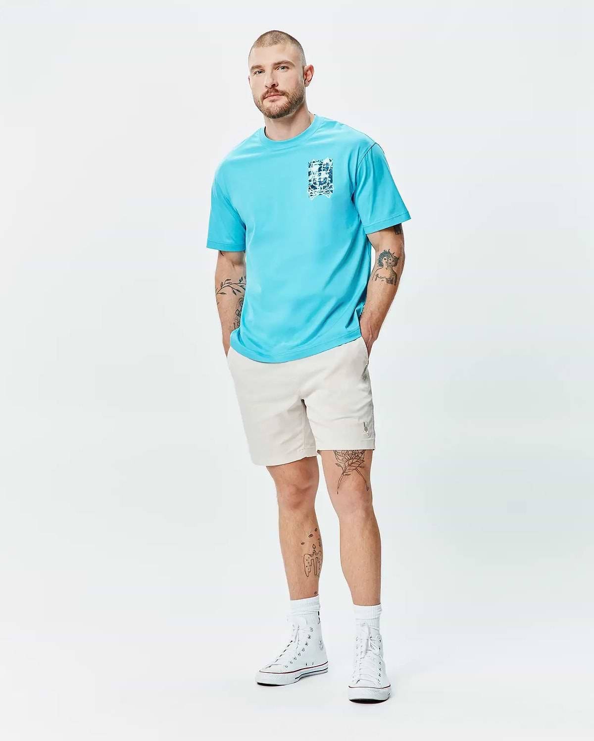 Mens Isaac Relaxed Fit Tee in aqua blue jersey, featuring a blue and white graphic print on the left chest, paired with light beige relaxed shorts with a small embroidered Psycho Bunny logo on the left thigh.