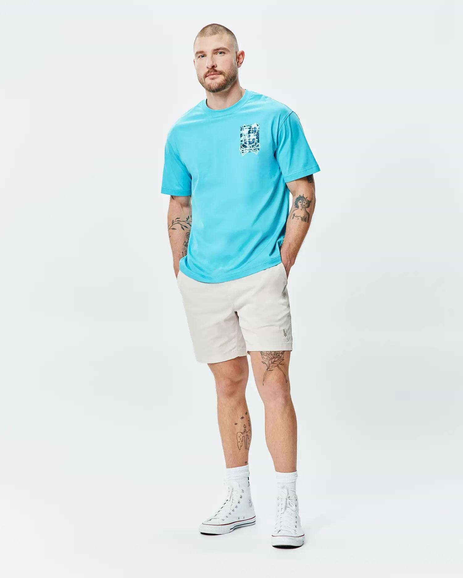 Mens Isaac Relaxed Fit Tee in aqua blue jersey, featuring a blue and white graphic print on the left chest, paired with light beige relaxed shorts with a small embroidered Psycho Bunny logo on the left thigh.