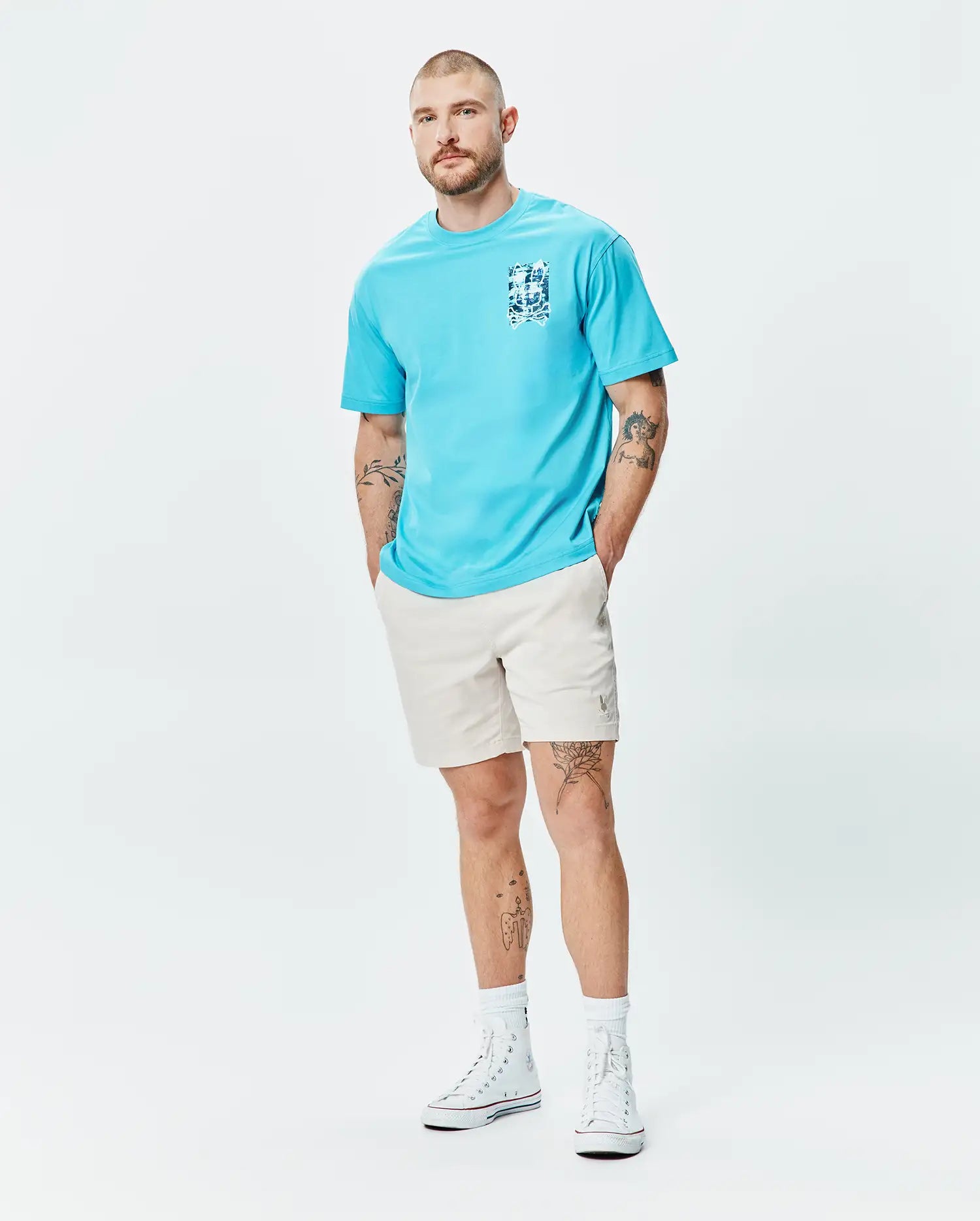Mens Isaac Relaxed Fit Tee in aqua blue jersey, featuring a blue and white graphic print on the left chest, paired with light beige relaxed shorts with a small embroidered Psycho Bunny logo on the left thigh.