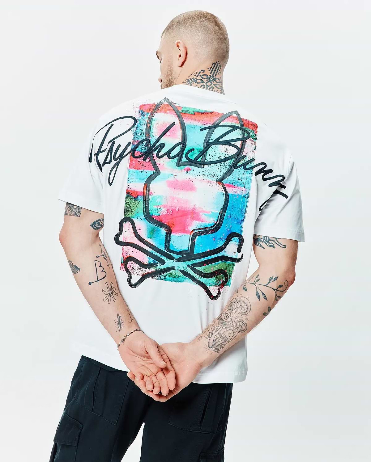 Mens Isaac Relaxed Fit Tee, a white soft jersey t-shirt with a large back graphic of a vibrant, watercolor-style Psycho Bunny skull logo in pink, blue, and green, overlaid with bold black script.