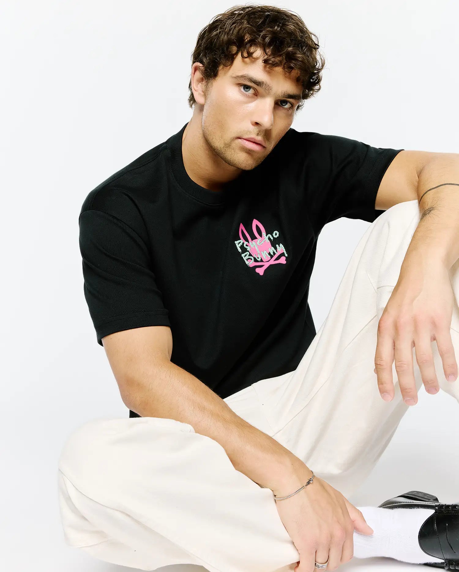 Mens Duncan Relaxed Fit Tee, a black jersey t-shirt with a relaxed fit, featuring a vibrant neon pink and light green Psycho Bunny skull logo graphic print on the chest.
