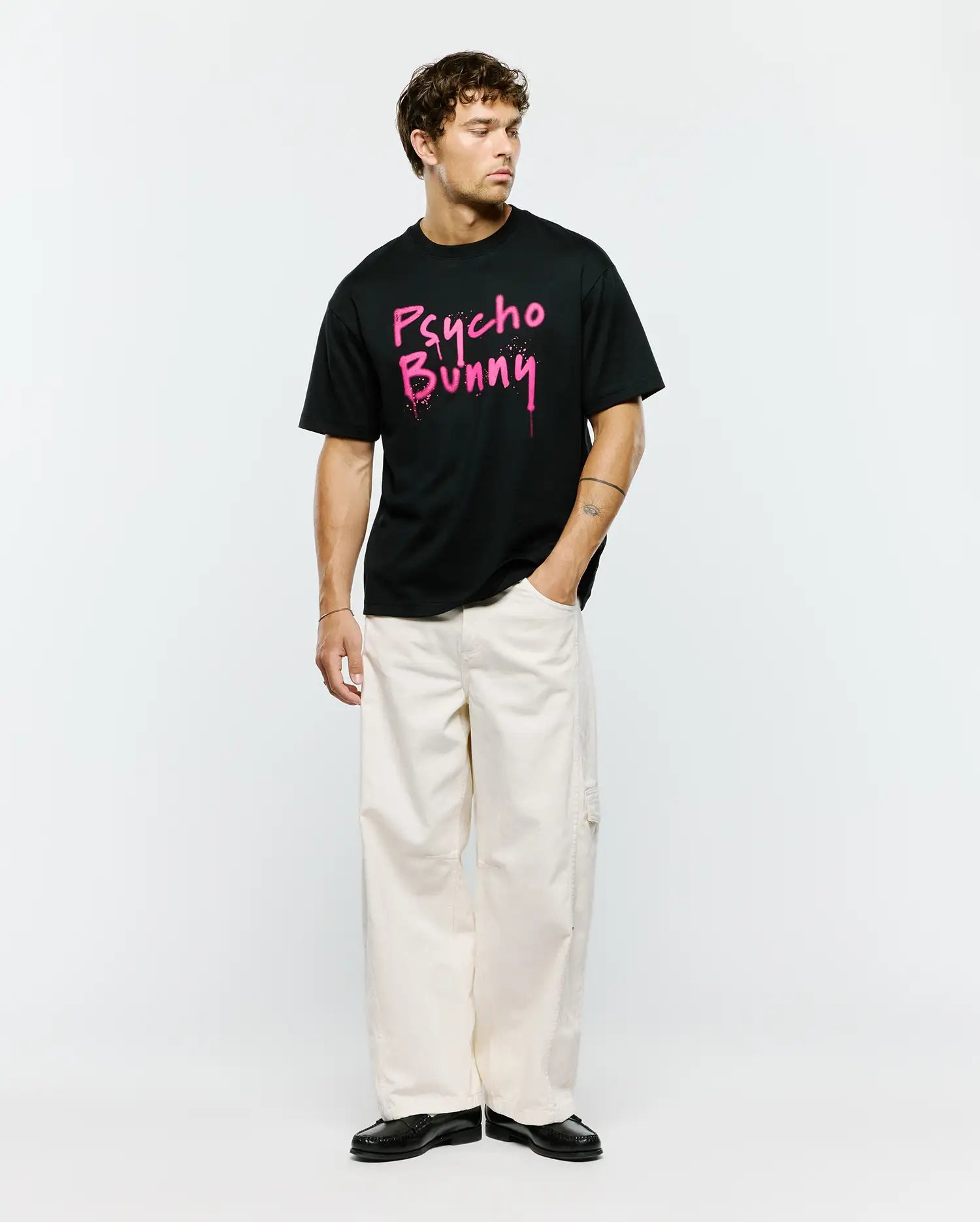 Mens Duncan Wordmark Relaxed Fit Tee, a black jersey t-shirt featuring a bold, large pink graffiti-style Psycho Bunny wordmark graphic with splatter details on the center chest.