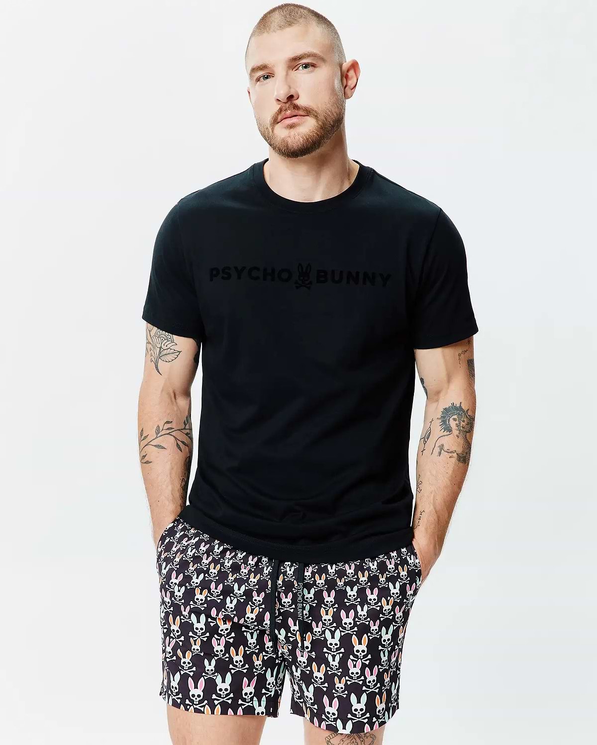 Mens Royce Graphic Tee, a black jersey t-shirt with a tone-on-tone PSYCHO BUNNY logo on the chest, paired with black shorts featuring a multi-colored bunny skull pattern. (2)