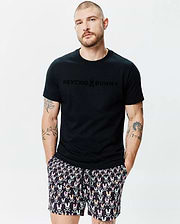 Mens Royce Graphic Tee, a black jersey t-shirt with a tone-on-tone PSYCHO BUNNY logo on the chest, paired with black shorts featuring a multi-colored bunny skull pattern. (2)